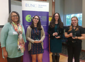 Four women, three holding trophies, stand in front of the Three Minute Thesis banner.