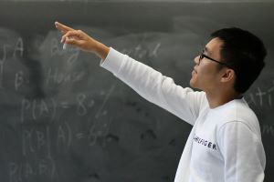 A graduate student in a white long-sleeved shirt points at a chalkboard.