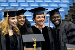 Four doctoral graduates in black regalia smile and pose together at the UNC Chapel Hill doctoral hooding ceremony.
